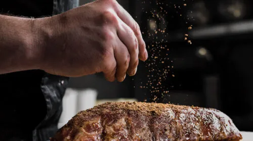 Raw piece of meat, beef ribs. The hand of a male chef puts salt and spices on a dark background, close-up.