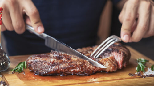 Hands holding fork and knife and eating delicious juicy steak on a wooden plate